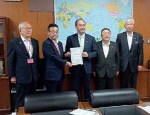 大きく引き延ばされた世界地図が貼られた壁の前で、要望書を一緒に持つ男性2人、両側に市長を含めた男性3人が横に並んでいる写真