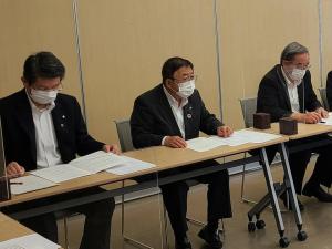 長机の席につき手元の資料を見ている様子の関係者の男性2名と、顔を正面に向けている市長の写真