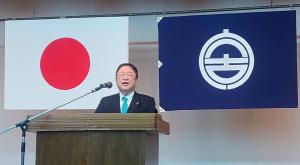 日の丸と市旗が壁に飾られ、その前の演台で挨拶をしている市長の写真