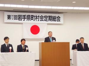 「第10回岩手県町村会定期総会」の横断幕が飾られ白色の胸章リボンをつけた男性2名が演台左に座り演台で市長が挨拶をし、右側にも参加者の方々が座っている定期総会の写真