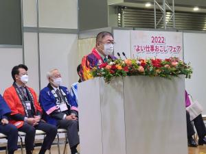 市長が後ろに法被を着た参加者が座っている、色とりどりの花で飾られた、演台で式辞を述べている写真