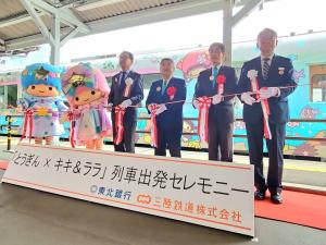 イメージキャラクターでラッピングされた電車が停車するホームで、テープカットをする様子の市長と3名の男性、キキとララの着ぐるみの写真