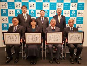 前列に賞状の額縁を持って椅子座る4名、後列に立つ3名の関係者が並んでいる記念写真