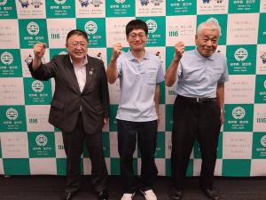 「第12回全国和牛能力共進会鹿児島大会」に参加する 高田泰義さん、高田耕蔵さんと市長がバックパネルの前で右腕を曲げ握った拳を顔の横に掲げているポーズを決めている記念写真
