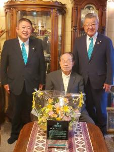 木澤健一先生が座っている机に花束と記念楯が置かれ、その両脇に市長と男性1名が立っている写真