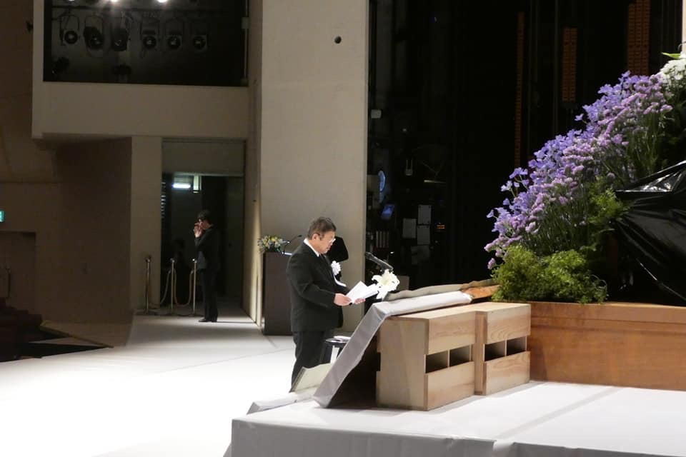 献花台の前で、式辞用紙を両手で広げスタンドマイクに向かい話をする様子の市長の写真