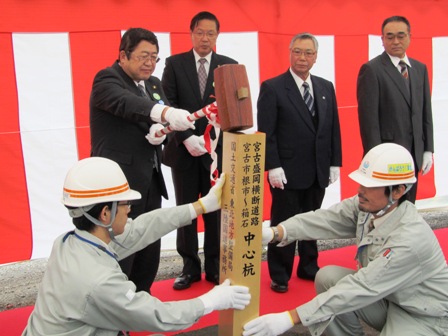 紅白幕の前に並ぶ関係者が見守る中、ヘルメットに作業着姿の男性二人が両側で支える中心杭を、市長がハンマーで杭を打つ写真