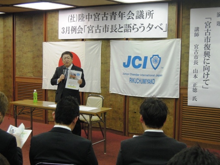 社団法人陸中宮古青年会議所3月例会「宮古市長と語らう夕べ」と書かれた横断幕の下に日本の国旗と国際青年会議所の社旗の前で市長が話している写真