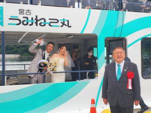宮古うみねこ丸に乗船しピースのポーズししているタキシードを着た男性、ウェディングドレスを着た女性と、船の前方で赤い胸章を着け佇む市長を写した写真