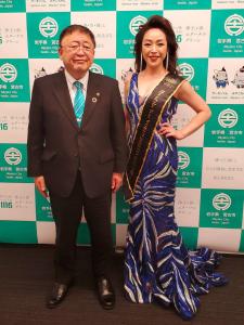 バックパネルの前で青のノースリーブドレスを着て、左手を腰にあててポーズを決める「ミセス・オブ・ザ・イヤー」に出場した佐藤和美さんと、隣に立つ市長の写真