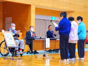 スタンドマイクの前に立つ3人の方が挨拶をし、車いすに座った男性や観覧の市長等が拍手をしている様子の写真