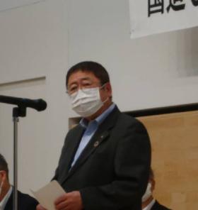 「国道340号宮古岩泉間整備促進期成同盟会」と書かれた横断幕が貼られた会場で、マイクスタンドの前で挨拶を行っている市長を右斜めの角度から撮影した写真