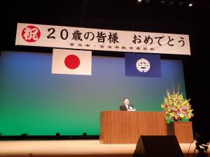 「祝 20歳の皆様 おめでとう」と書かれた横断幕があり、舞台の演壇に立つ市長の写真
