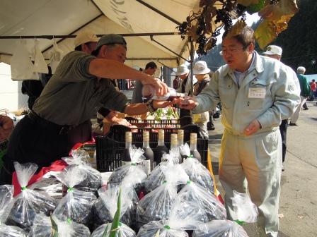 グレーの作業服姿の山本市長が、出店された方からヤマブドウのジュースを試飲用にコップに注がれている写真