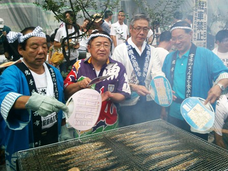 はっぴ姿の市長と関係者が、網の上で焼かれたさんまをうちわで仰いでいる写真