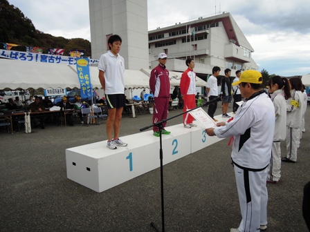 同じ高さの数字が続いた台に男性6名の入賞者たちが並び市長が表彰状を読み上げている表彰式の写真