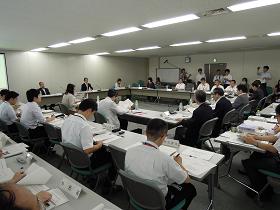 会議室にロの字型で並べられた長机の席に関係者が座っており、机の上にある資料を読んでいる様子の写真