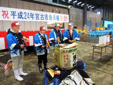 「祝平成24年宮古市魚市場初売り」と書かれた横断幕の前で青色の半被を着た男性4名が酒樽を叩いている写真