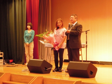 スピーカーやキーボードが置かれたステージ上で、花束を持つ八神純子さんと市長がマイクを持ち話をする様子の写真