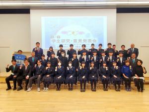 学生研究意見発表会と映し出されたスクリーンを背景に、市長の他関係者の方々、発表した高校生の皆さんがガッツポーズをとった姿で3列に並んで撮影した集合写真