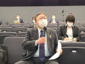 椅子が設置された会場で、椅子に座り右手にマイクを持って話している様子の市長と、後方の椅子に座る数名の関係者の写真