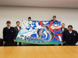 茶色いテーブルの後方で、大漁旗を持って広げる学生4名、関係者の男性1名と市長の写真