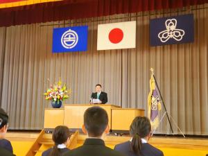 宮古市の市旗、国旗、校旗が掲揚されたステージ上の演台で、市長が新入生に向けて祝辞を述べている写真