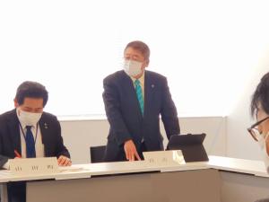 役員会で立ちあがって意見を述べている市長と隣でメモをしている男性の写真