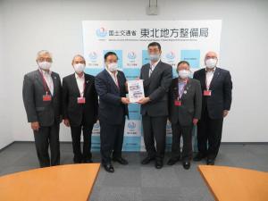 「国土交通省 東北地方整備局」と書かれたバックパネルの前で、6名のスーツを着た男性が横一列に並び、中央の2名が要望書を一緒に持ち記念撮影をしている写真