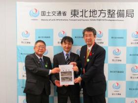 国土交通省東北地方整備局と書かれたボードの前で、市長と山田町の佐藤信逸町長、梅野修一局長の3人で整備促進に関する要望書を持っている写真