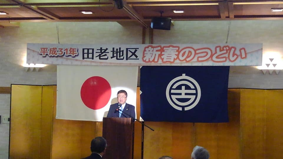 2枚の旗と「平成31年田老地区新春のつどい」と書かれた横断幕に金屏風が設置された会場で、スポットライトをあび壇上に立つ市長が挨拶を行っている様子の写真