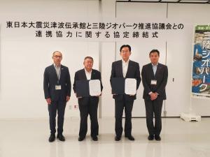 協定締結式の横断幕が貼られた会場の前にスーツ姿の男性4名が並び、中央の2名が締結所を持っている写真