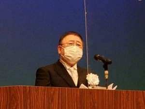 白い胸章を着け、演台の前で話をする様子の市長の写真