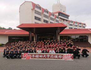 「歓迎 早稲田大学応援部」と書かれた看板が地面に置かれ、その後方にしゃがんで何列にもなって並ぶ市長や大勢の学生たちの記念写真