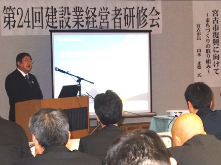 「第24回建設業経営者研究会」の横断幕の下「宮古市復興に向けて」と題した資料がスクリーンに映し出されており、演壇で講演をする市長と、その話を聞く参加者の写真