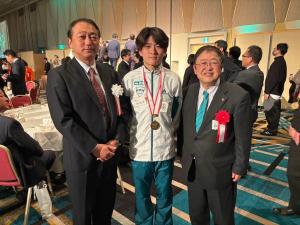 白色の胸章リボンを付けたスーツ姿の男性と赤色の胸章リボンを付けた市長の間に青山学院大学陸上競技部の選手が並んで3人で記念撮影をしている写真