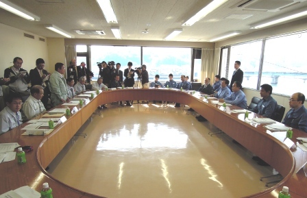 平野達男復興対策担当大臣と大臣に同行されている方々、宮古市職員が楕円形状に座り、市長が自席で立って、報道陣のスタッフがいる中、復興計画案や地区のまちづくりについて報告している写真