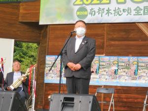 両手を身体の前で組みスタンドマイクの前で話をする市長と、その斜め後方で椅子に座りトロフィーを手に持っている男性の写真