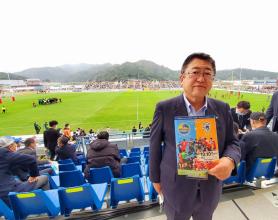 芝生のグラウンド場と観客席を背景に市長が試合のパンフレットを持って、客席通路に立っている写真