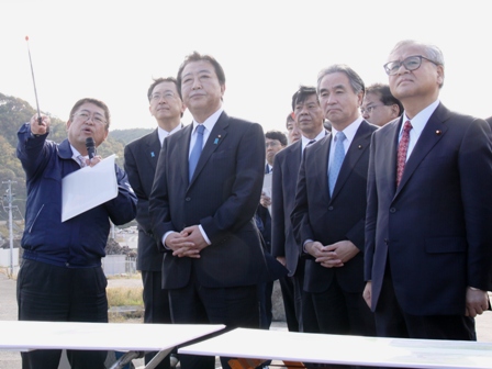 柵のような前で、市長がマイクを持ち指示棒を使って、野田佳彦首相と関係者らに説明をする様子の写真