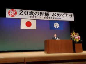 「祝 20歳の皆様おめでとう」と書かれた横断幕が飾られ舞台の演台横にはきれいな花が飾られ市長が挨拶をしている写真