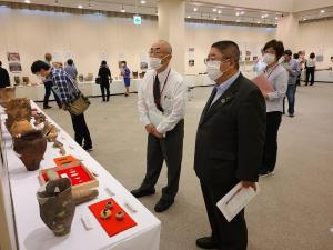 会場の壁際に沿って並べられた机の上に、発掘された出土品が並べられ、それを鑑賞している来場者の方々と市長、説明をしている様子の関係者の男性の写真