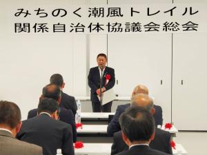「みちのく潮風トレイル 関係自治体協議会総会」と書かれた横断幕が吊るされた会場で、赤い胸章を着けてスタンドマイクに向かい話をする様子の市長と、並べられたテーブルに2名ずつ座り話を聞く様子の関係者の方々の写真