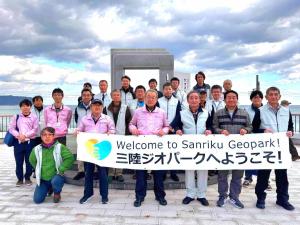 海をバックに「三陸ジオパークへようこそ!」と書かれた横断幕を持った市長と参加者が3列に並んでいる集合写真
