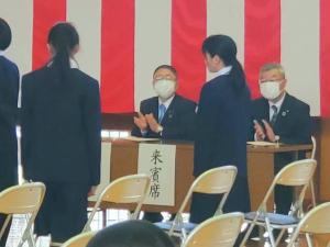 壁に紅白幕が設置された体育館で「来賓席」と書かれた紙が貼られた席に座った男性と市長が、新入生入場時に拍手している入学式の様子の写真