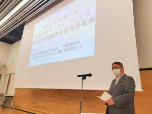 画面が映し出されたスクリーンの斜め前方で、スタンドマイクの前で資料を手に持ち話をする様子の市長