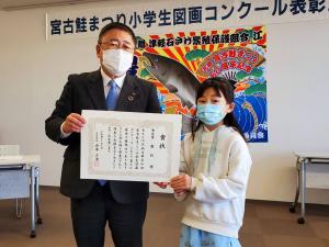 「宮古鮭まつり小学生図画コンクール表彰式」と書かれた横断幕と鮭の絵が書かれたポスターのようなものが貼られた会場で、一緒に賞状を持ち正面に向ける市長と女子小学生が写った写真