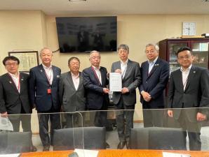テーブルの上にパーティションが設置された室内で、中央に国土交通省の堀田治港湾局長と市長会の代表が一緒に要望書を持って中央に立ち、市長を含む5名の関係者が横一列に並んでいる写真