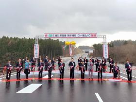 開通を祝う看板の下で割れたくす玉を背景に、道路に敷かれたレッドカーペット上で市長、他11人でテープカットをしている写真