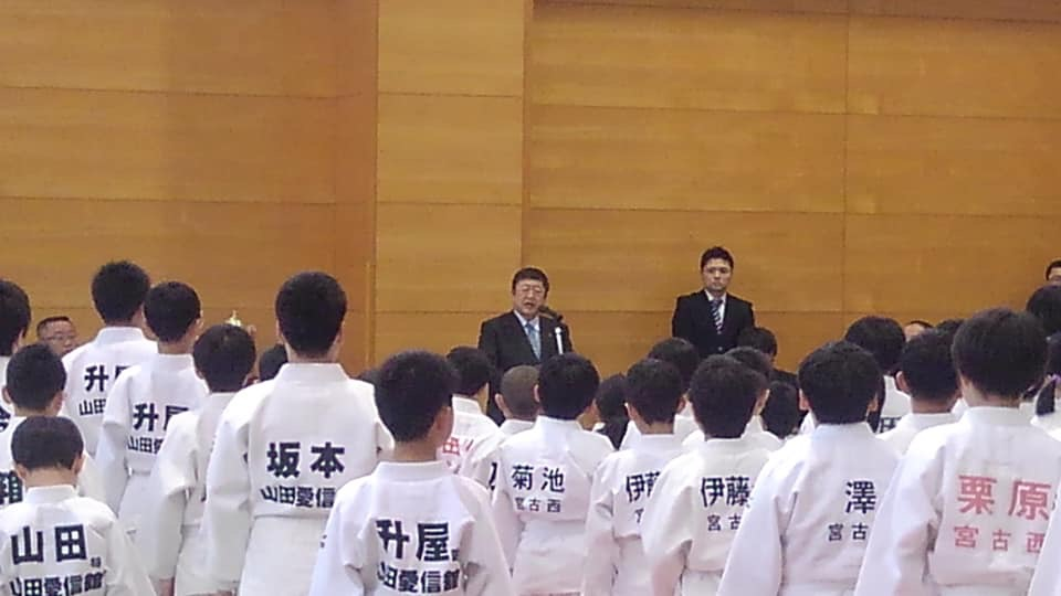 名前と所属先が背中に書かれた柔道着を着る少年たちが整列する前でスタンドマイクに向かい話をする様子の市長の写真